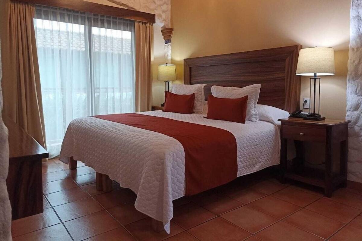 Una habitación de hotel con una cama grande, dos lámparas de noche, un cabecero de madera y una ventana con cortinas.