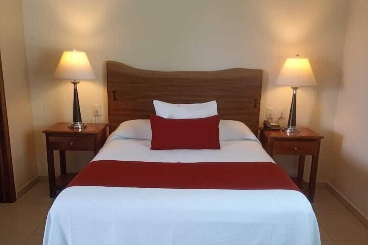 Una cama bien hecha con cabecera de madera, dos lámparas de noche y un almohadón rojo.