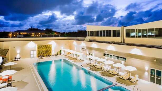 A swimming pool with sun loungers and umbrellas, surrounded by a building with large windows and a tiled floor. - Golden Tulip Le Diplomate Cotonou (Cotonou)