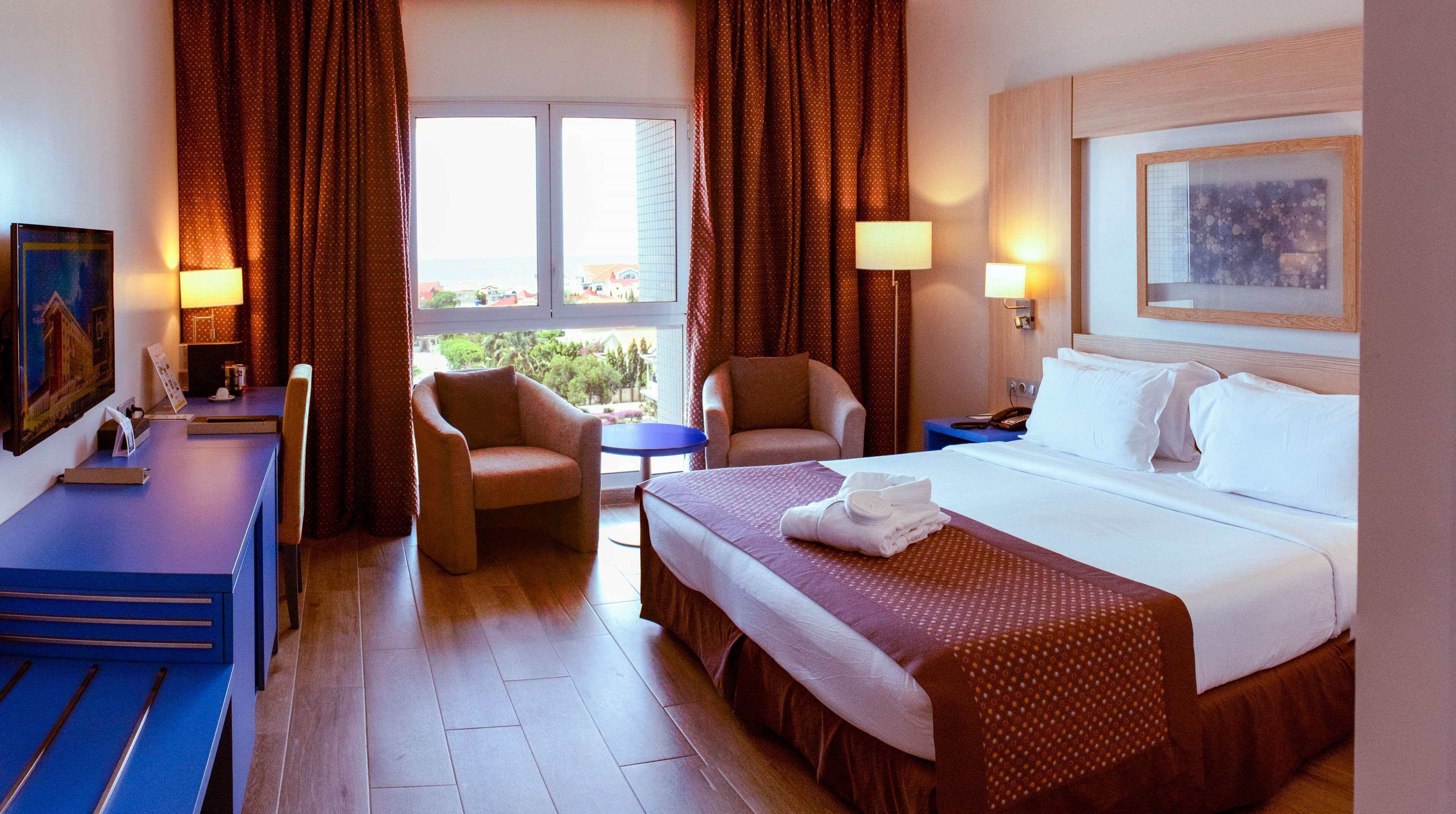 Una habitación de hotel con una cama, dos sillones, una mesita, un escritorio y un cuadro en la pared.