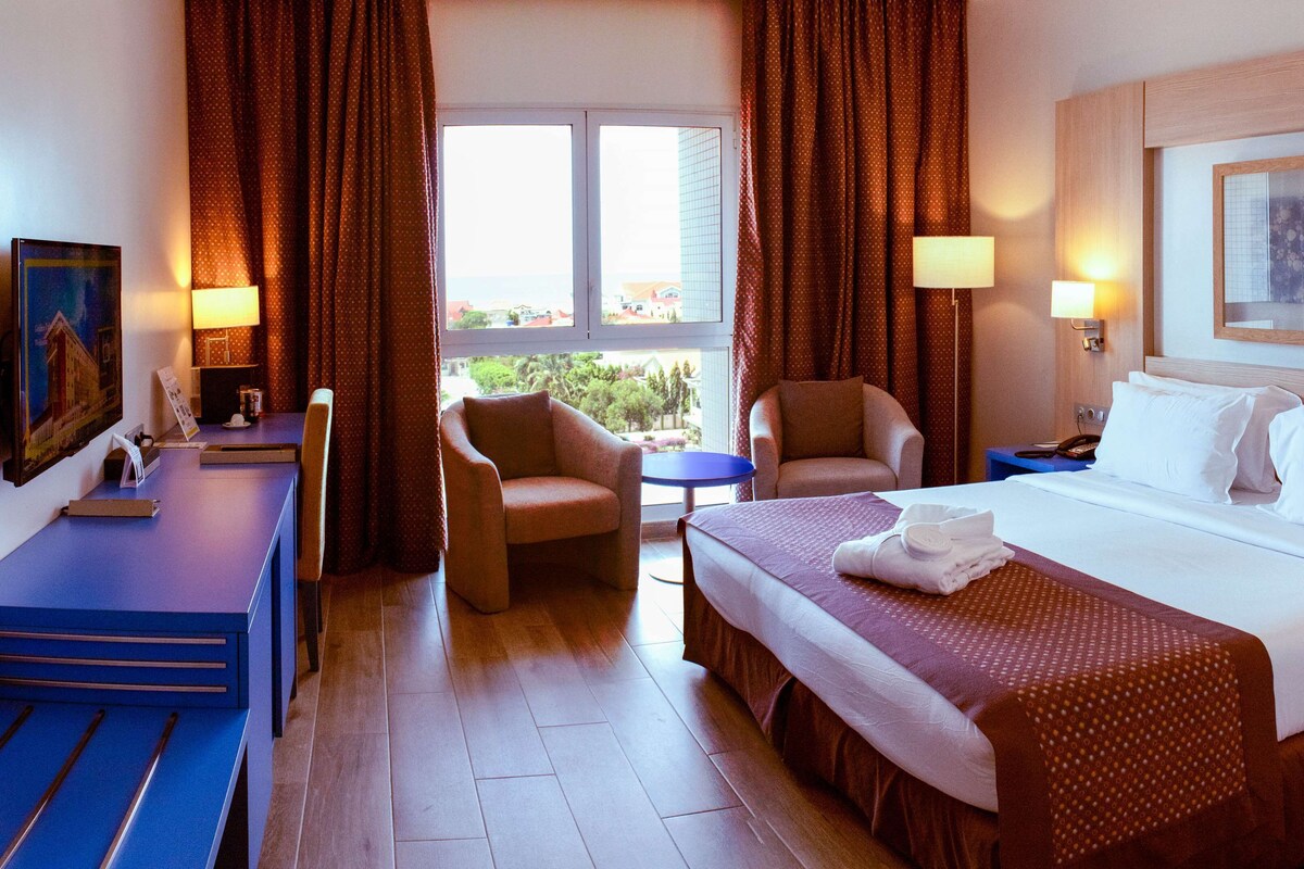Una habitación de hotel con una cama, dos sillones, una mesita, un escritorio y un cuadro en la pared.