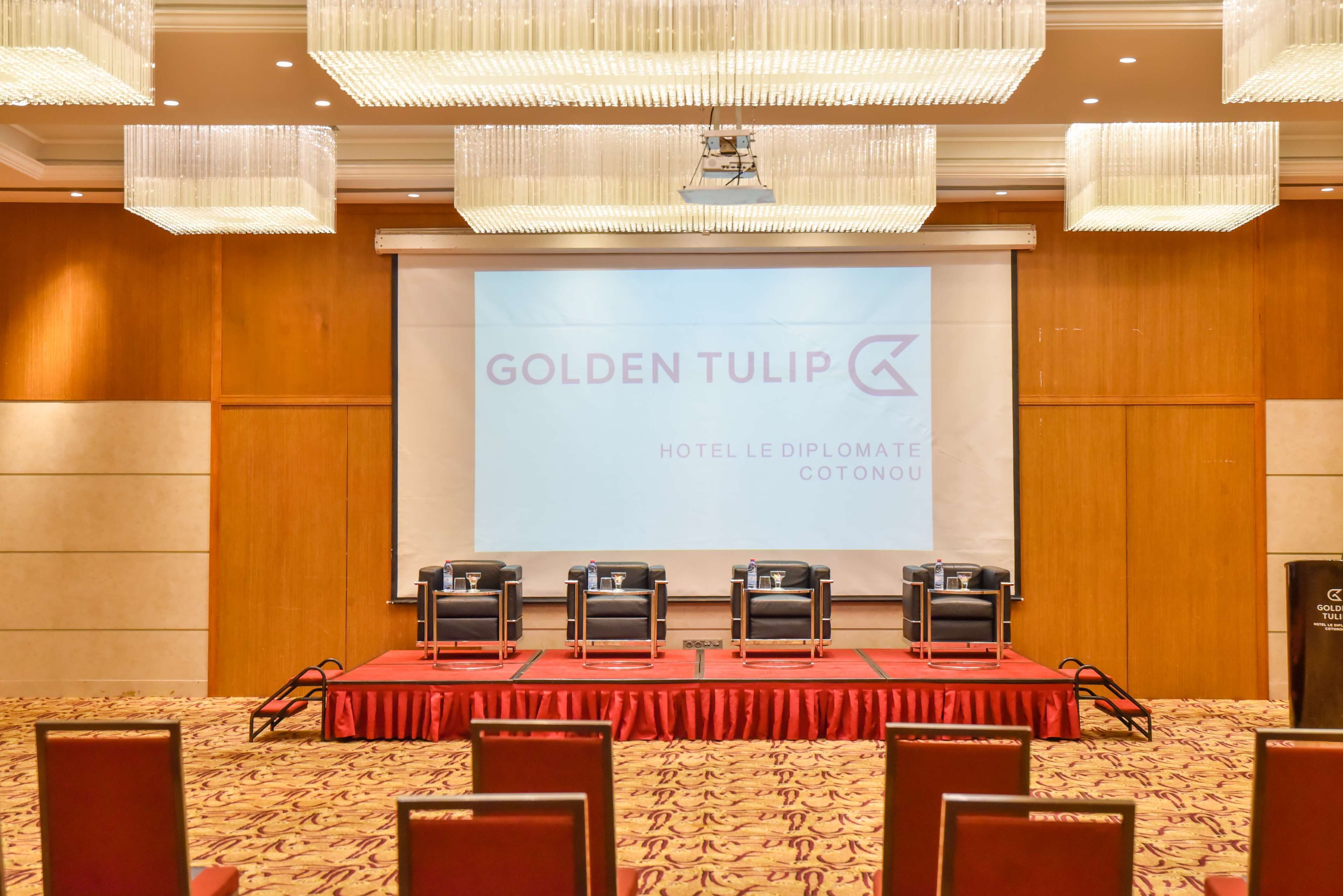 A conference room with a red table, black chairs, and a screen displaying "GOLDENTULIP HOTEL LE DIPLOMA COTONOU".