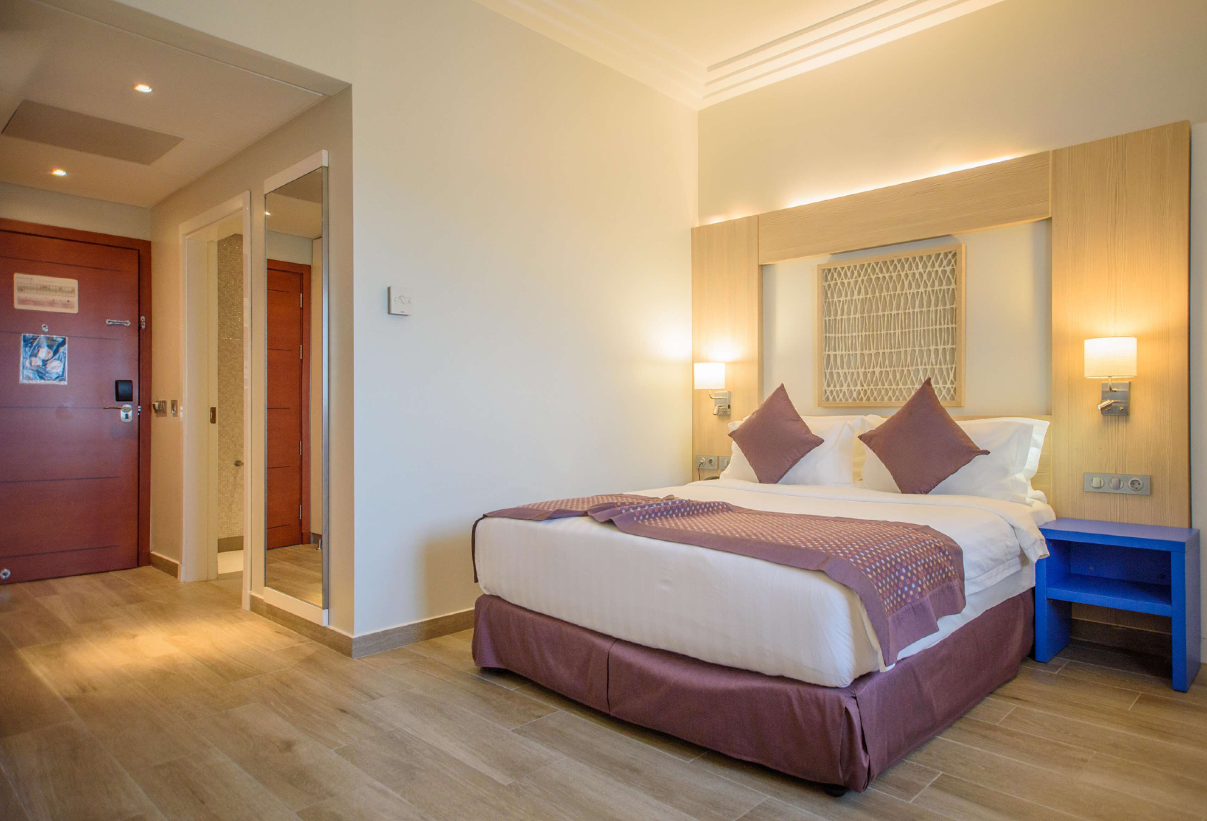 A hotel room with a large bed, a wooden door, a blue nightstand, and a wall-mounted light fixture.