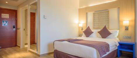 A hotel room with a large bed, a wooden door, a blue nightstand, and a wall-mounted light fixture.
