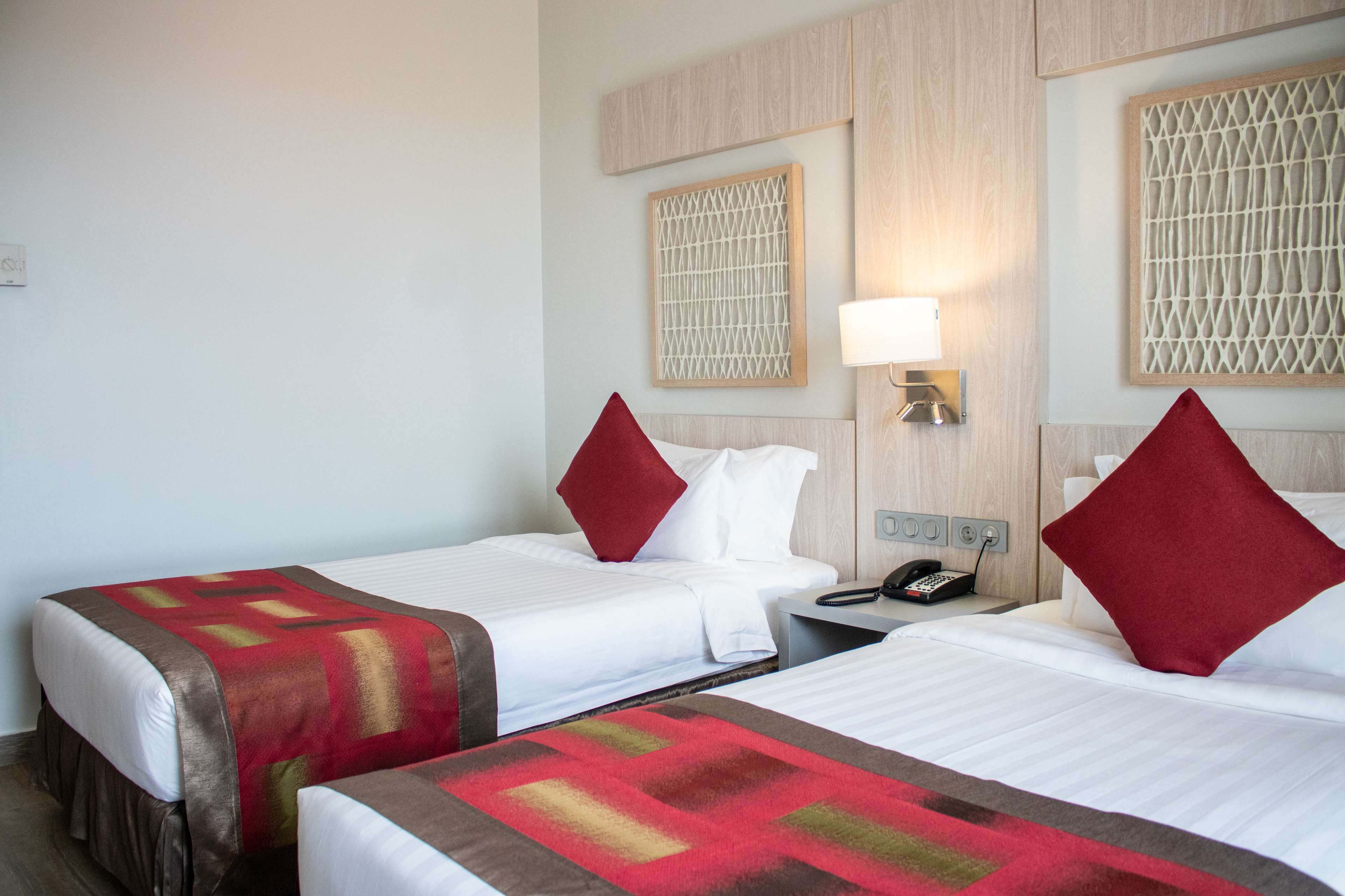 Ein Hotelzimmer mit zwei Betten, jeweils mit einem roten Kopfkissen, weißer Bettwäsche und einer roten und braunen Decke. Ein Nachttisch mit Telefon und Lampe ist vorhanden.