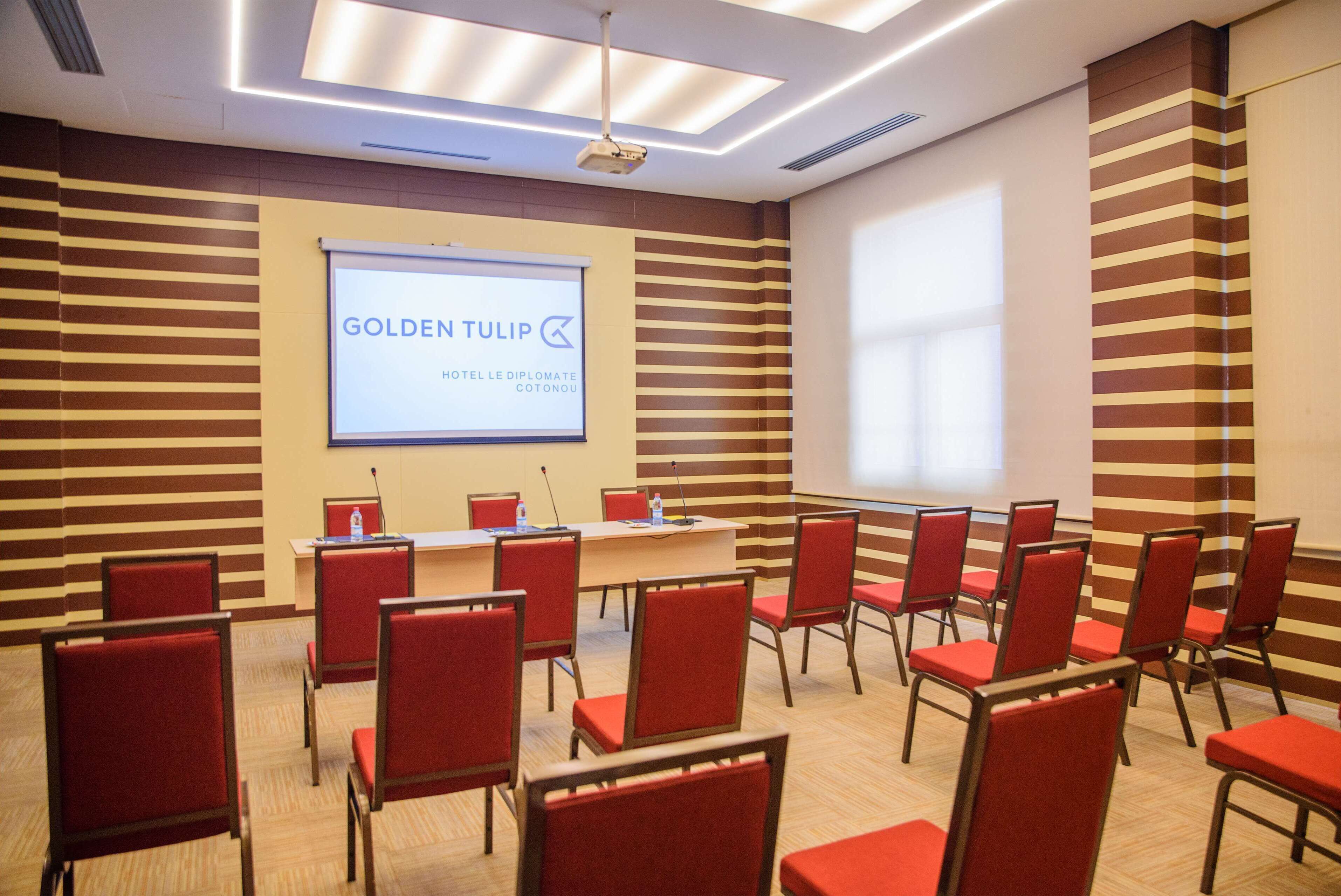 Un salón de conferencias con sillas rojas, una pantalla de proyector mostrando "GOLDEN TULIP HOTEL LE DIPLOMATIQUE COTONOU" y una mesa de madera con botellas de agua.