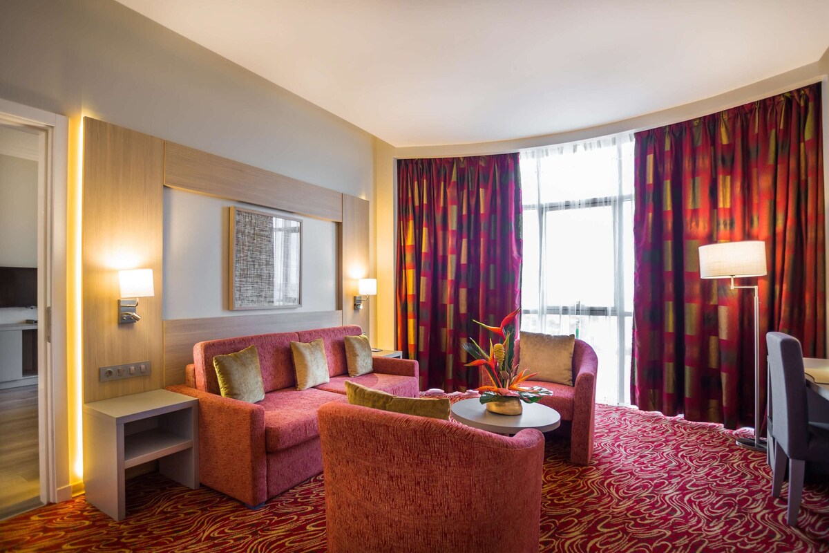 Una habitación de hotel con alfombra roja, un sofá, una silla, una mesita y una lámpara.