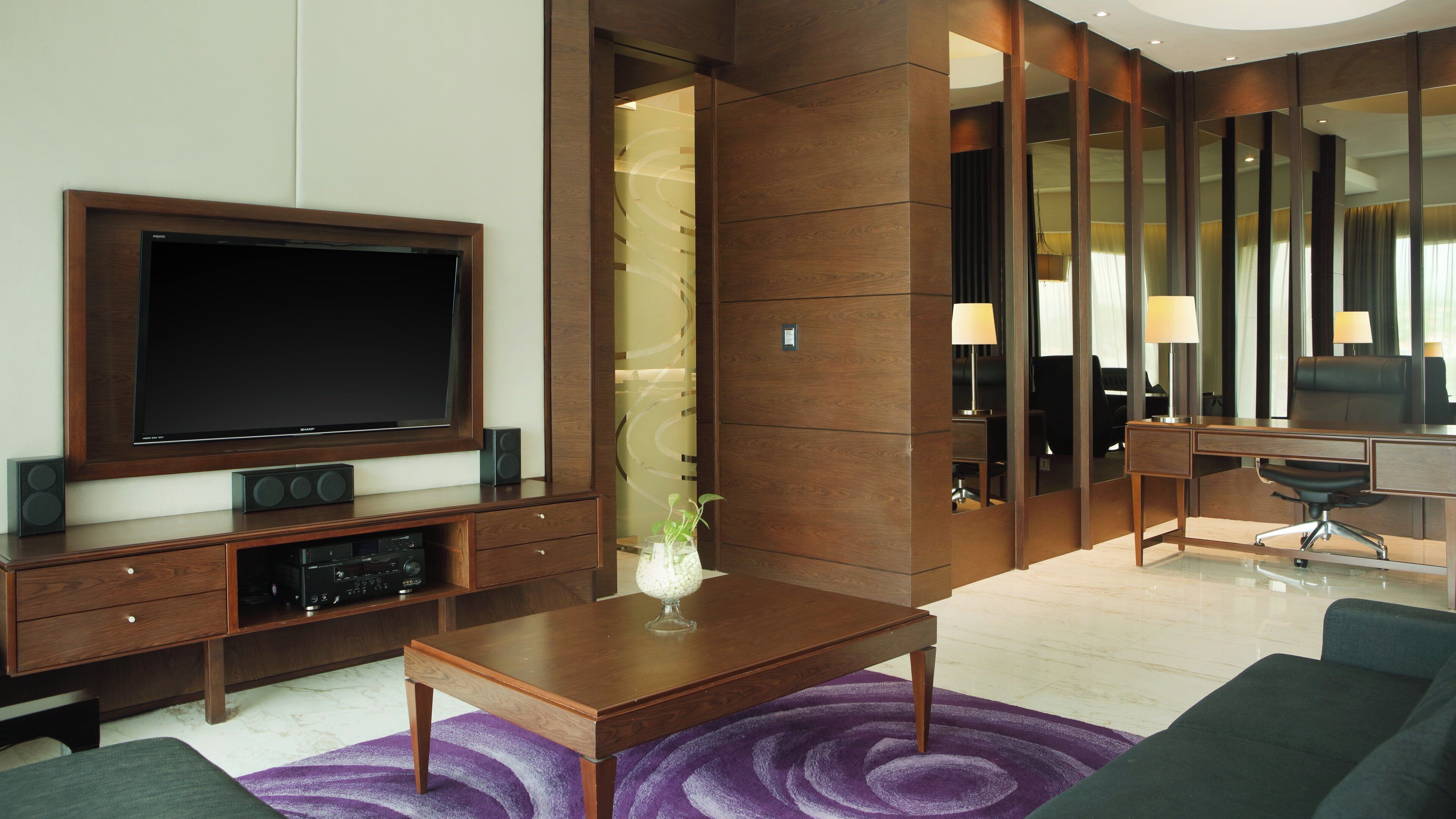A modern hotel room with a large flat-screen TV, a wooden desk, a comfortable sofa, and a purple patterned rug.