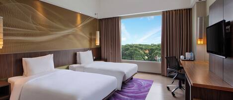 Quarto de hotel com duas camas, uma escrivaninha, uma televisão e uma janela grande com vista para árvores.