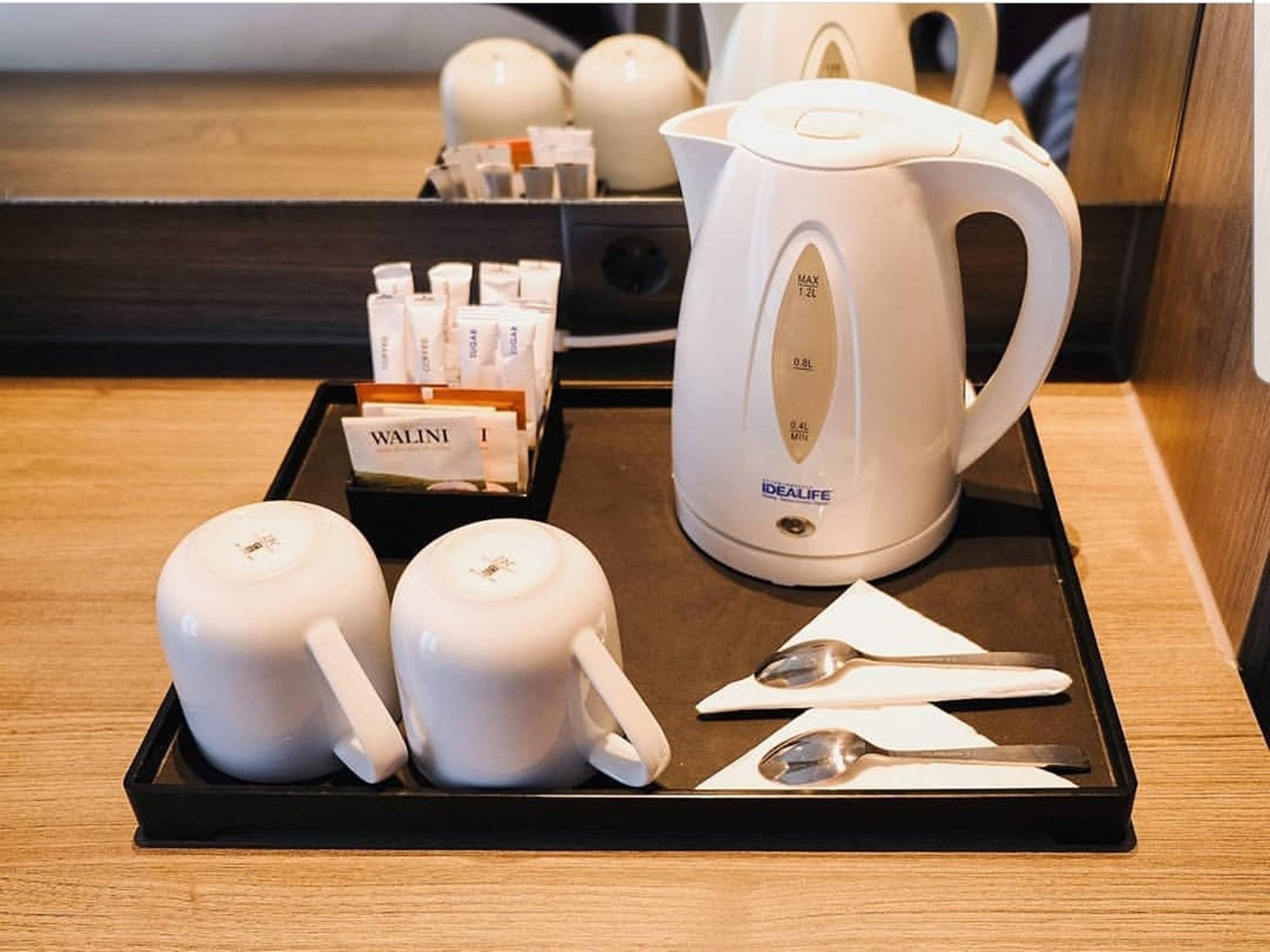 A tray with a white electric kettle, two white mugs, a box of coffee sachets, and two spoons on a wooden surface.