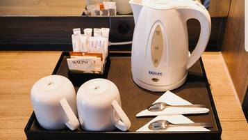 A tray with a white electric kettle, two white mugs, a box of coffee sachets, and two spoons on a wooden surface.