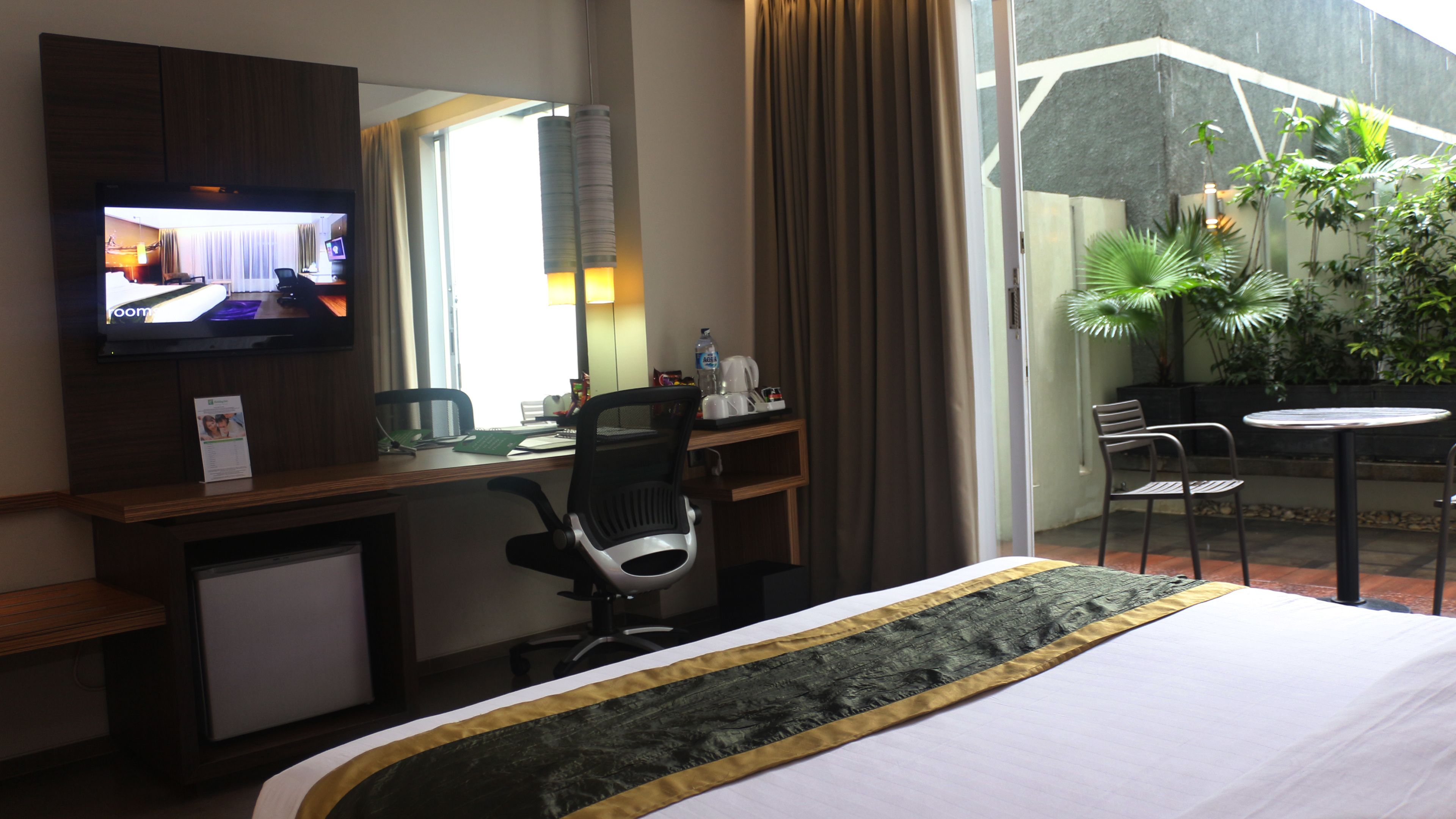 A hotel room with a bed, a desk with a computer, a television, and a small refrigerator. There is a window with curtains, a chair, and a small table with a plant.