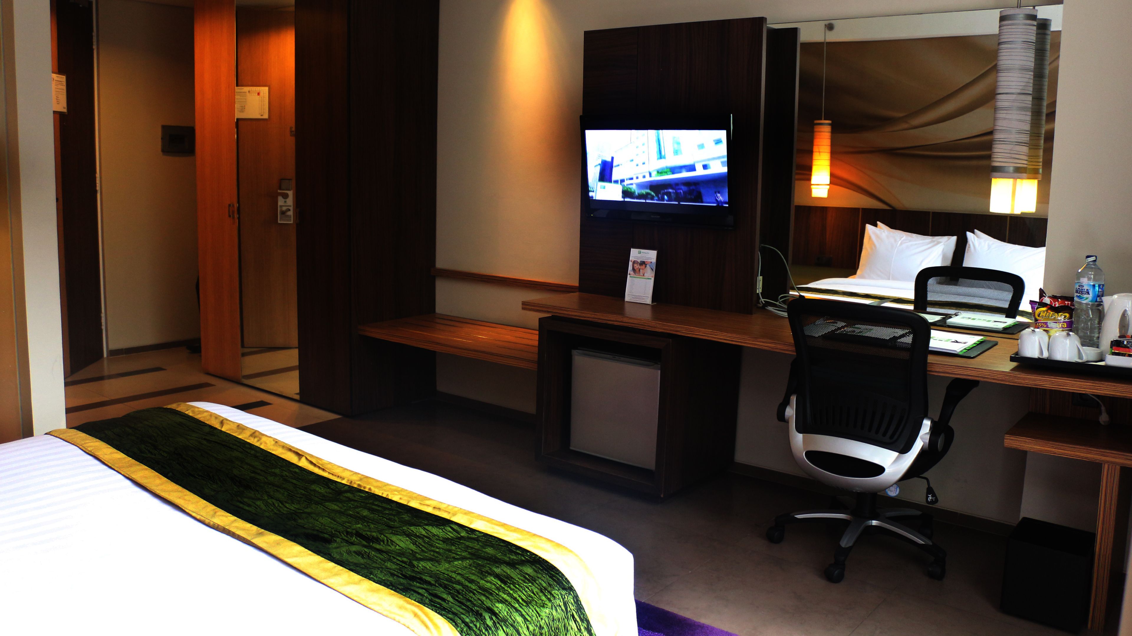 A hotel room with a bed, a desk with a computer, a chair, a television, and a small table with a bottle and a cup.