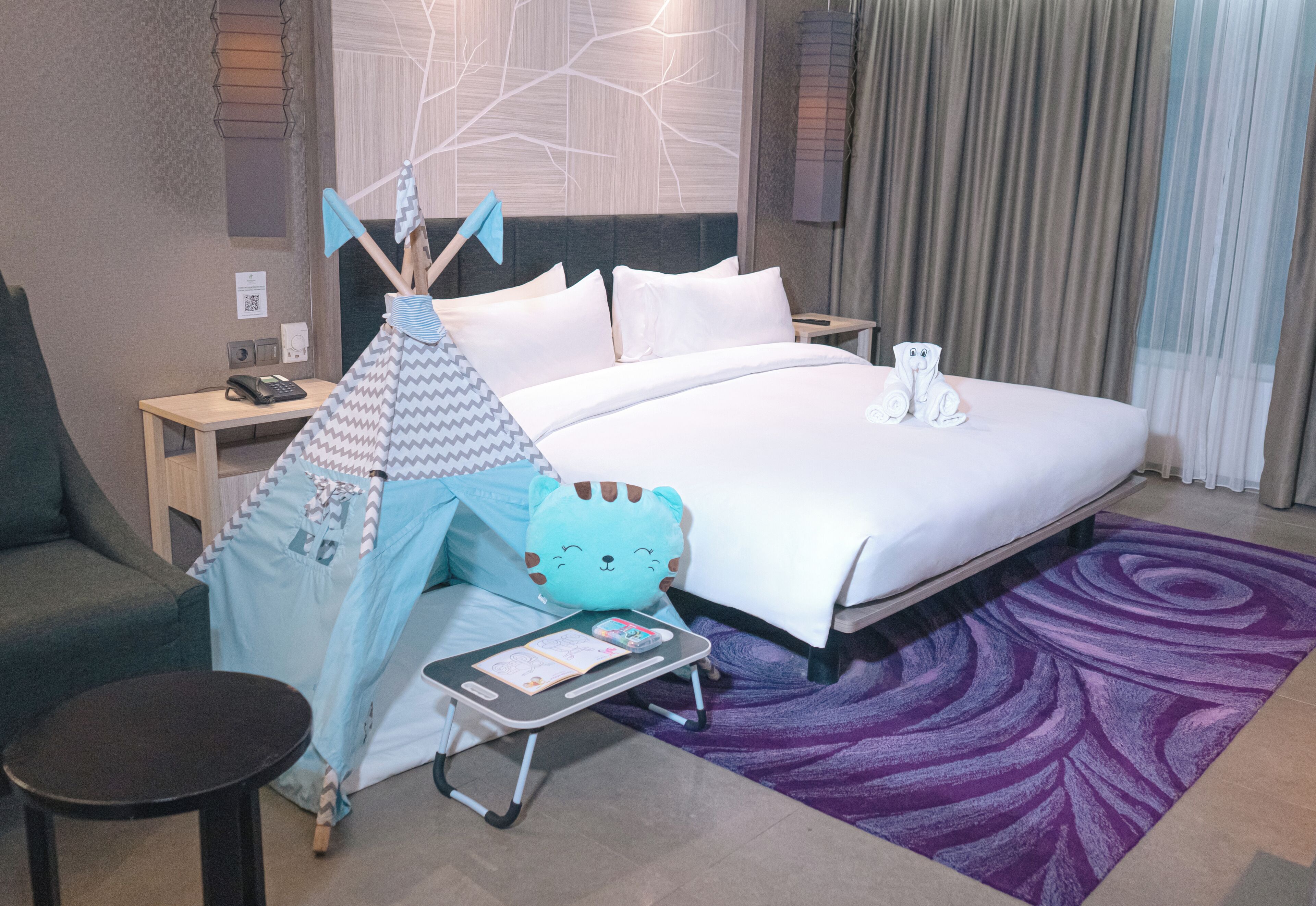 A hotel room with a bed, a small table with a phone, a chair, a tent, and a purple rug.