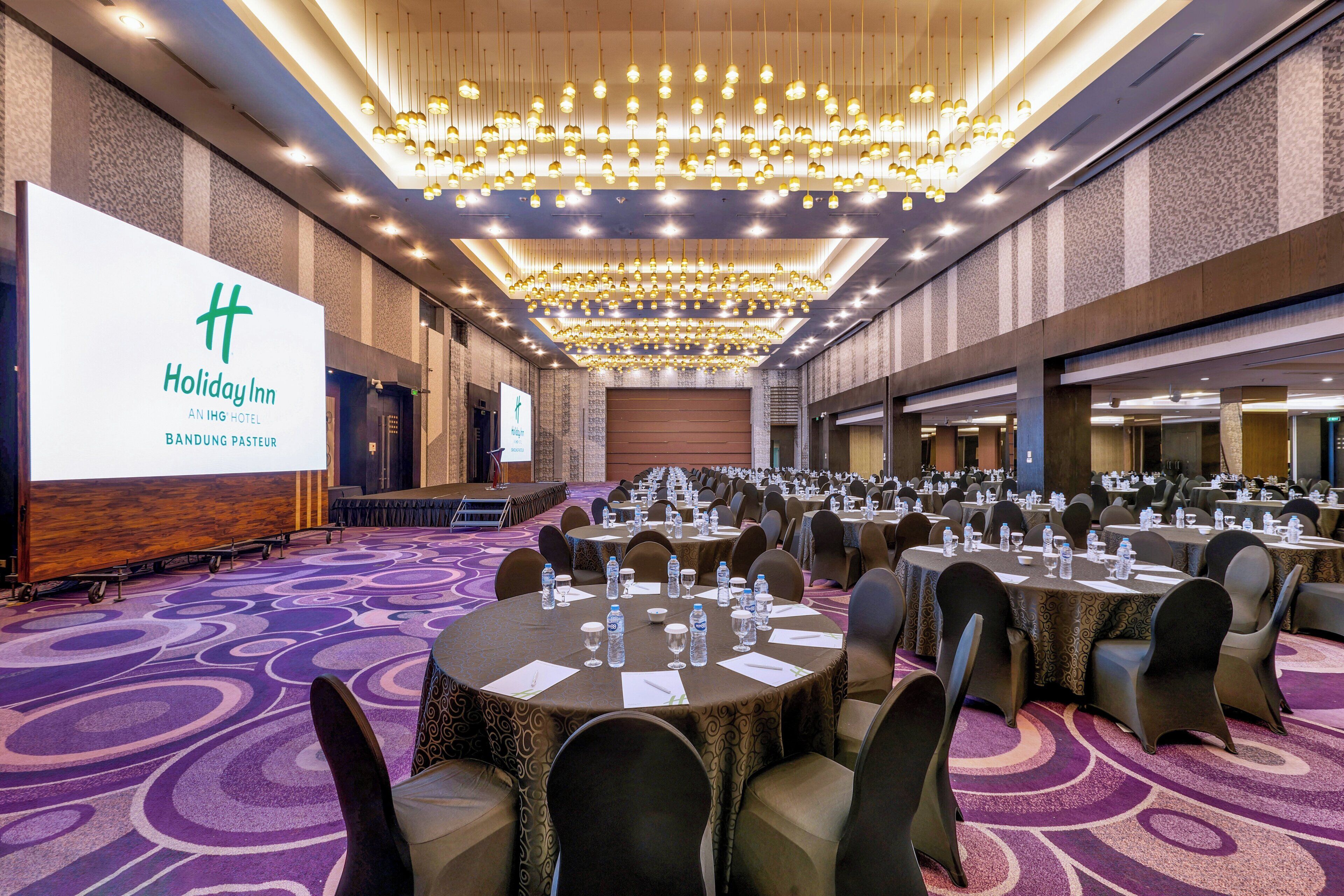 A large conference room with round tables, chairs, and a stage, featuring a prominent hotel sign.