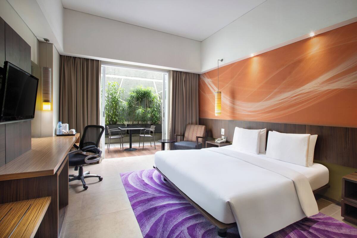 Ett hotellrum med en stor säng, ett skrivbord, en stol, en TV och utsikt över grönområden utanför.
