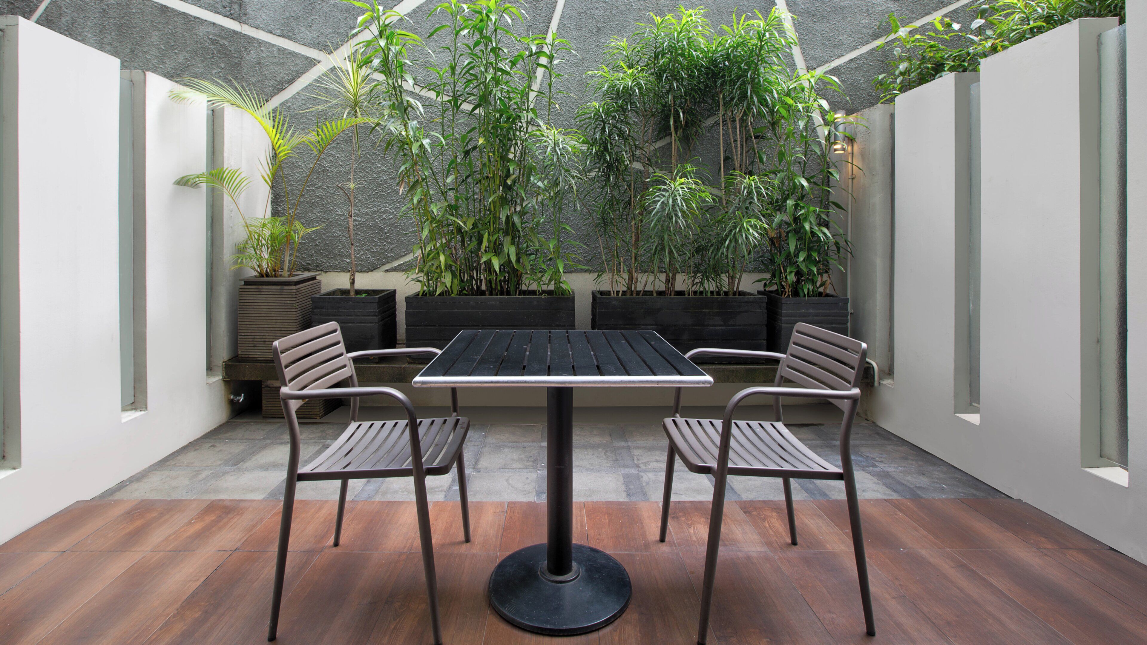 Un patio con una mesa y sillas, rodeado de macetas con plantas y una pared con textura.
