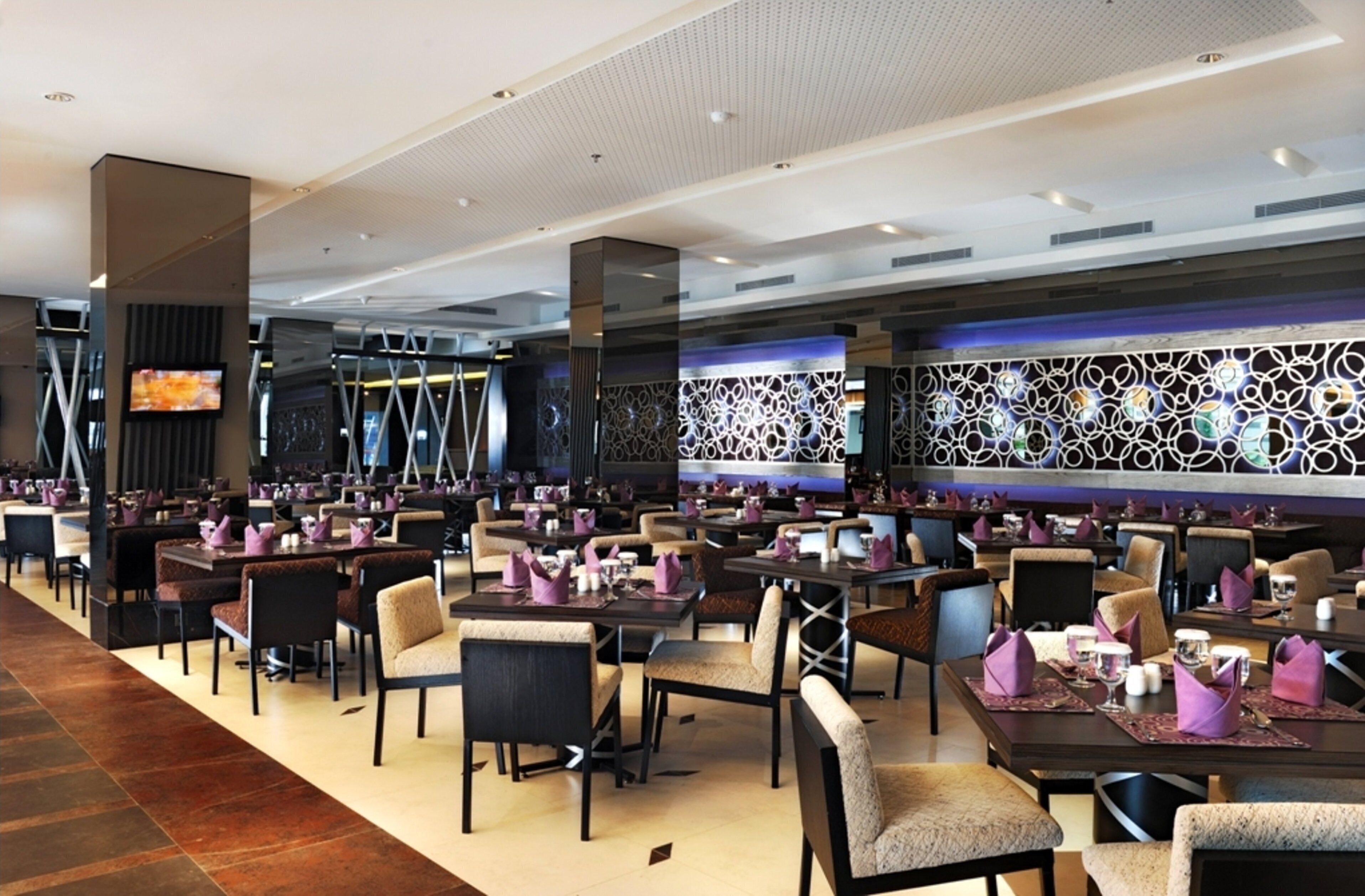 Un restaurante moderno con varias zonas de comedor, cada una con mesas y sillas, y una pared decorativa con un diseño geométrico.