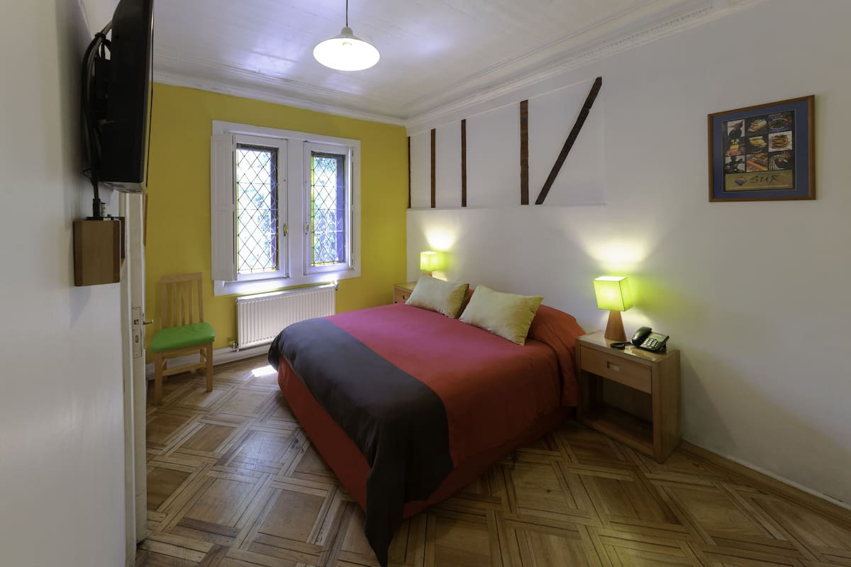 Un dormitorio con cama, lámparas de noche, ventana, una silla y un televisor montado en la pared.
