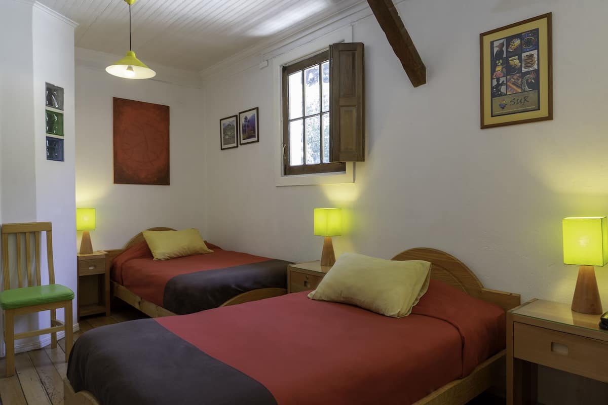 Una habitación con dos camas, una silla de madera, un ventanal y un cuadro en la pared.
