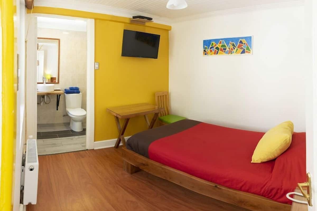 Una habitación con cama, televisor, un cuadro y un baño visible a través de una puerta abierta.