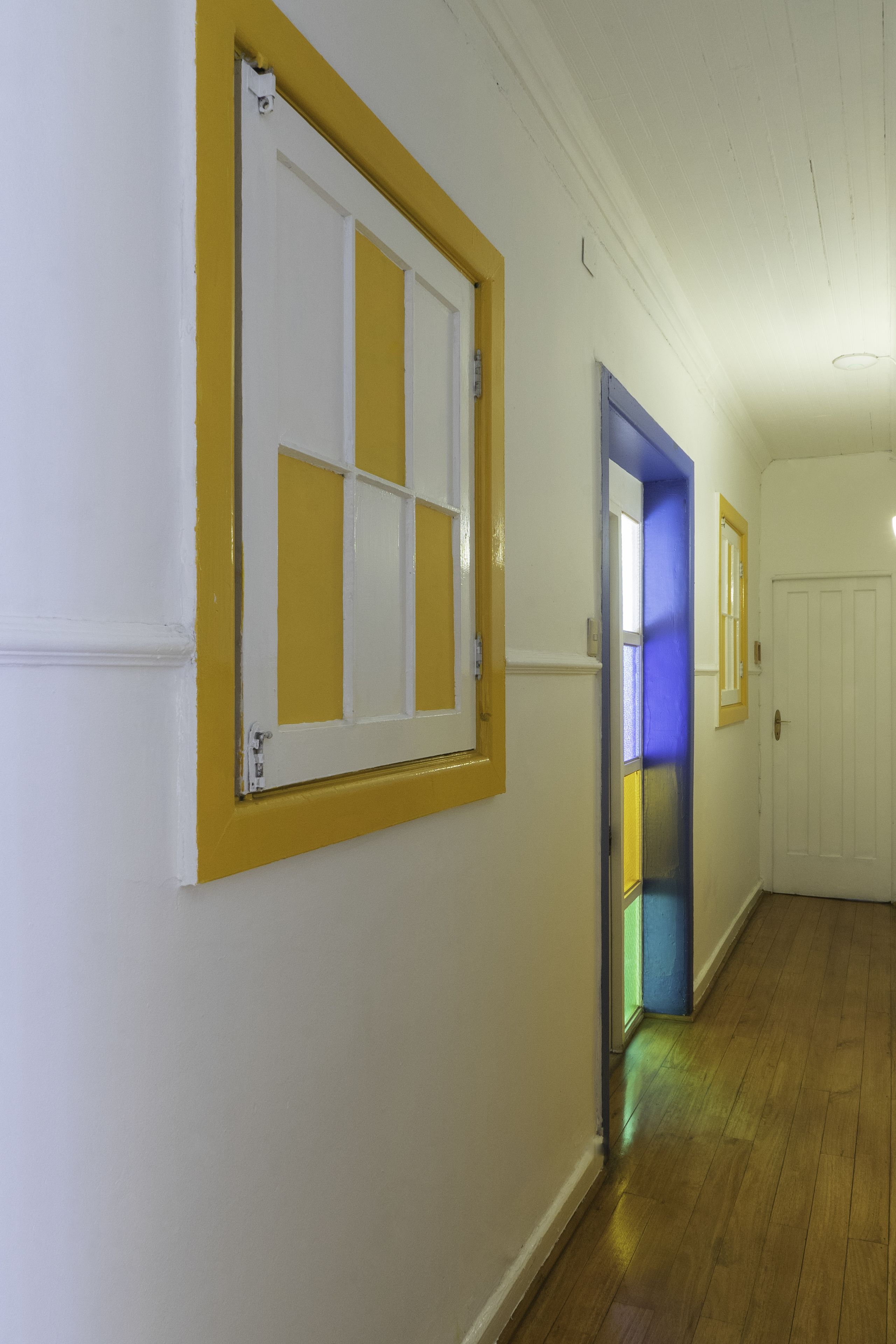 Un couloir avec une porte blanche, une fenêtre à cadre jaune et une porte à cadre bleu.