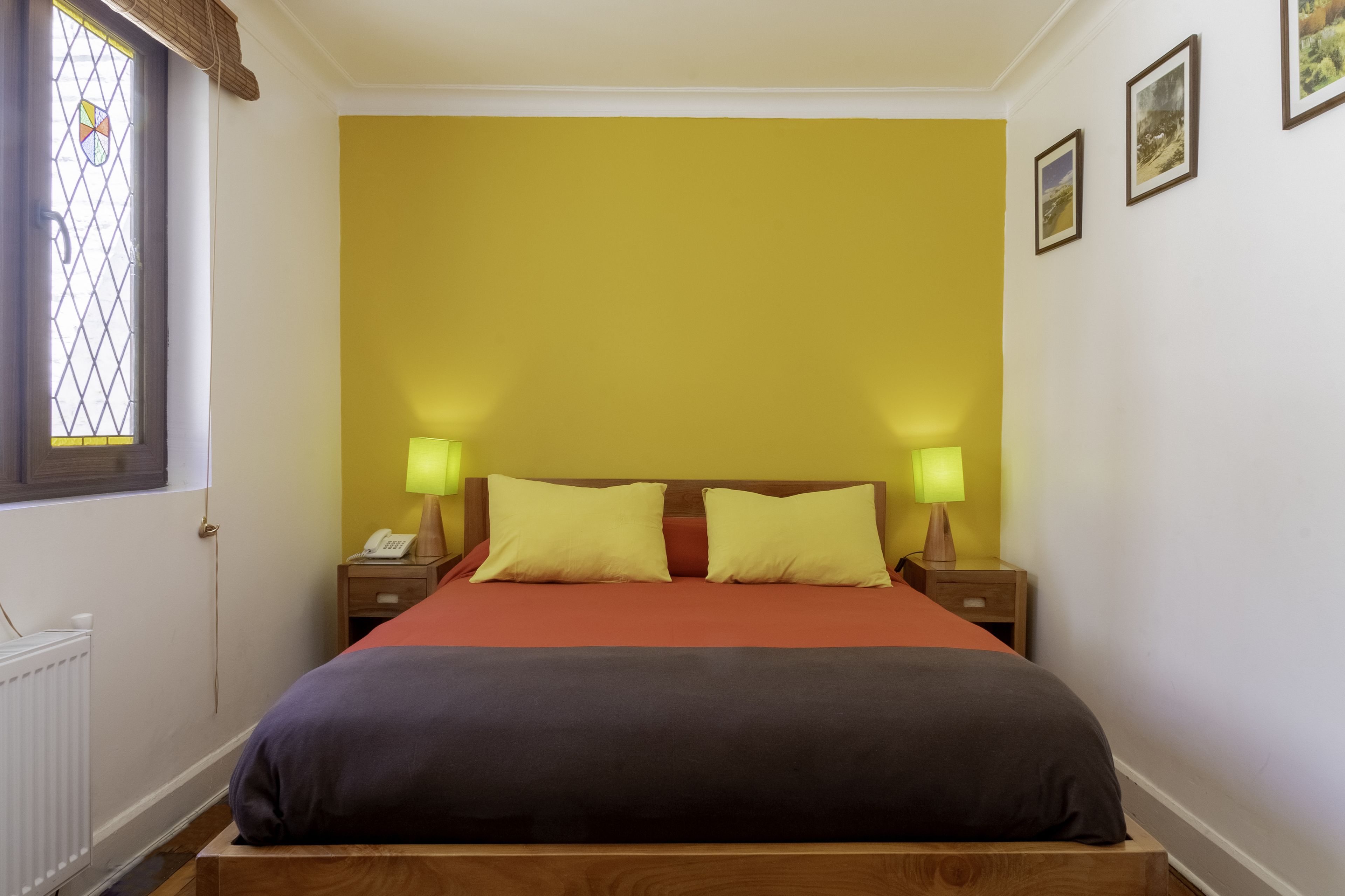 Un dormitorio con cama, dos mesitas de noche, una mesita auxiliar, un ventanal con diseño de vitral y una pared decorada con cuadros.