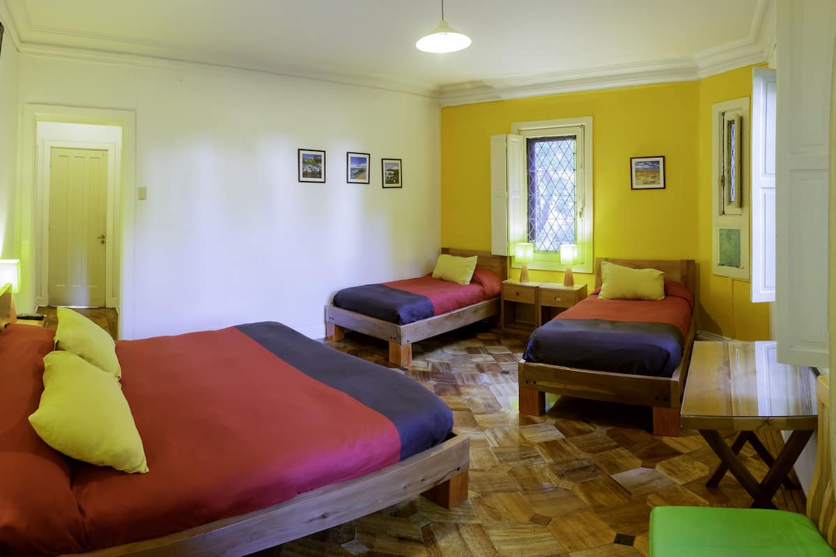 Una habitación con tres camas de madera, cada una con ropa de cama colorida, una mesita y una ventana con contraventanas.