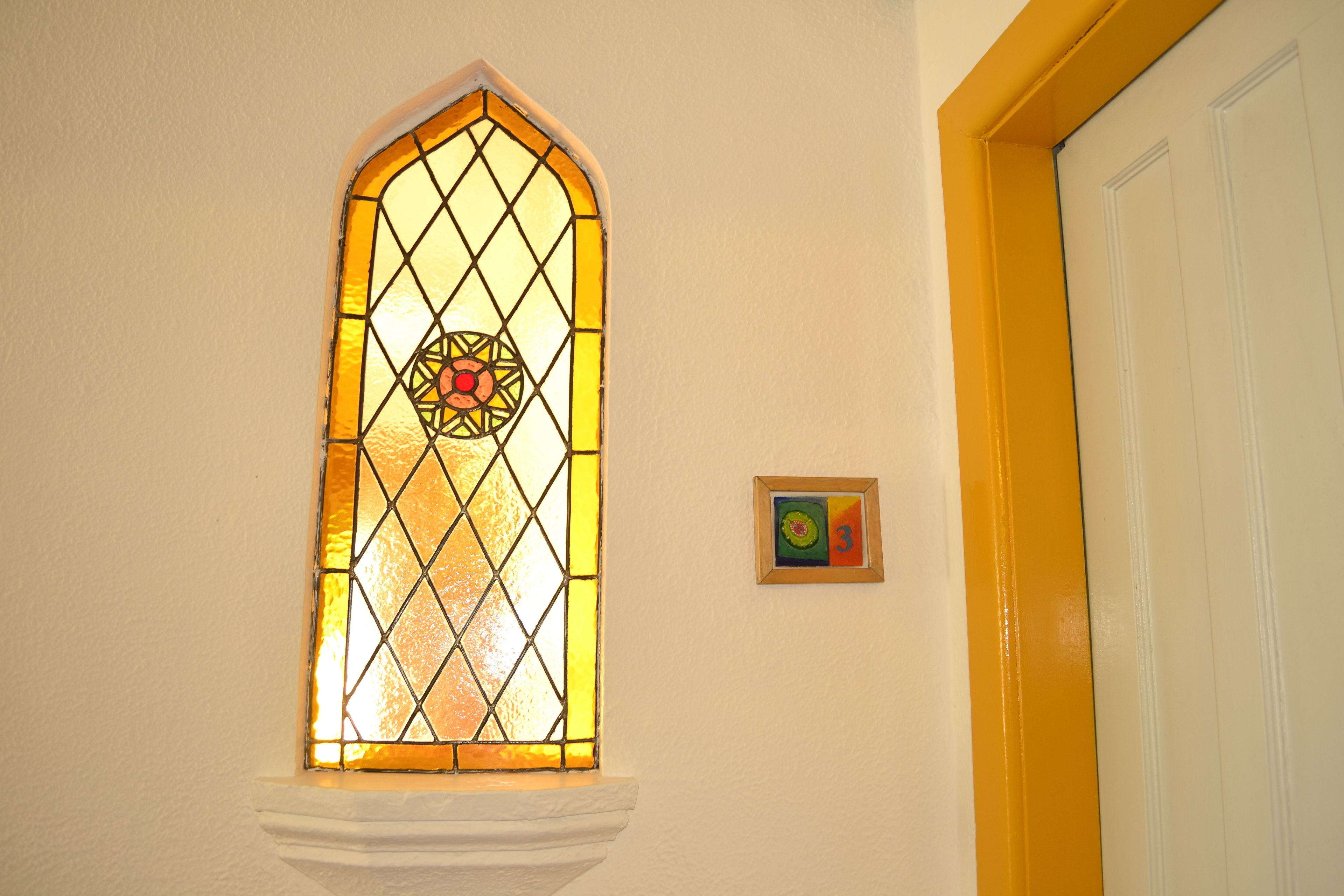 Une fenêtre en vitrail ornée de motifs géométriques, un chambranle de porte blanc et un petit tableau encadré accroché au mur.