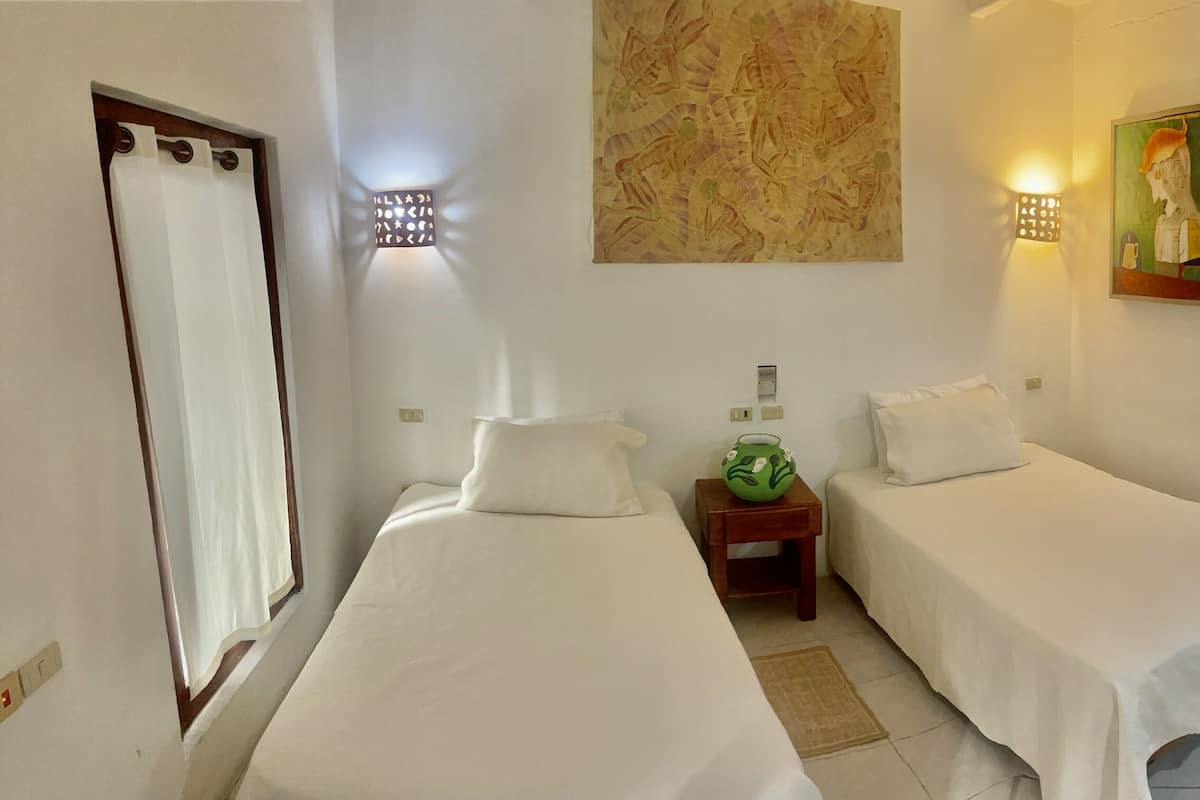 Habitación de hotel con dos camas individuales, una mesita con un adorno verde, una lámpara de pared y un cuadro.