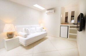 A minimalist white room with a sofa, a bedside table with a lamp, a small cabinet, and a mounted TV.