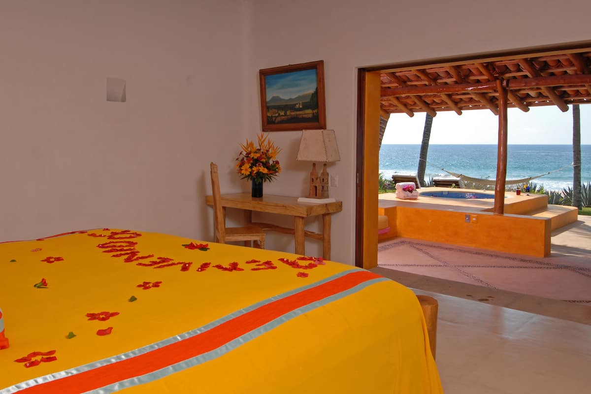 A room with a bed, a desk, a chair, a vase with flowers, and a view of the ocean.