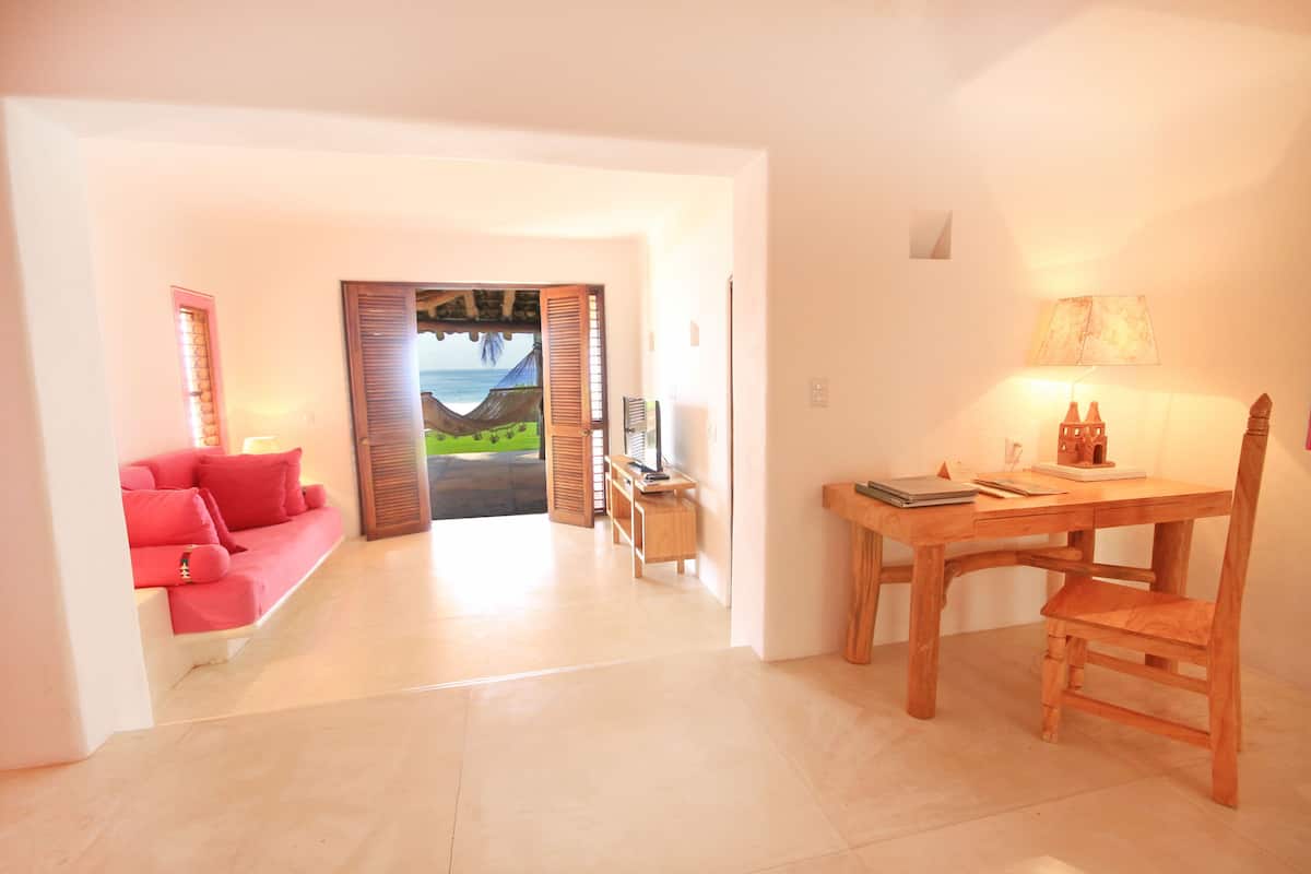 A spacious living area with a red sofa, a wooden dining table, and a view of a tropical landscape through an open door.