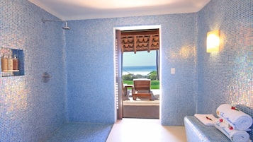 Un baño con mosaicos azules, ducha, banco y vista al mar a través de un vano abierto.