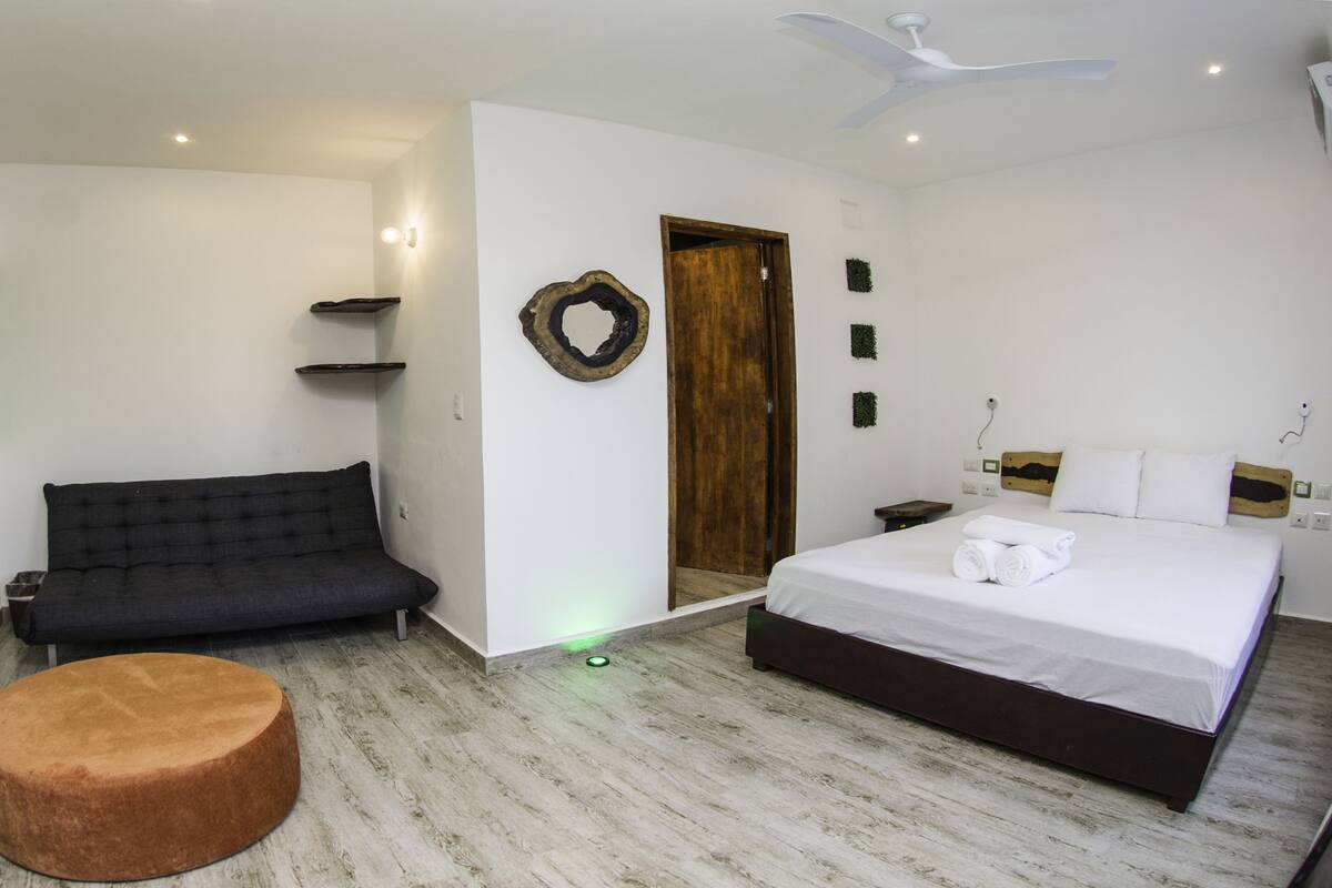Ein modernes Hotelzimmer mit einer Couch, einem runden Hocker, einem Bett mit weißen Leinen, einer Holztür und einem wandmontierten Fernseher.