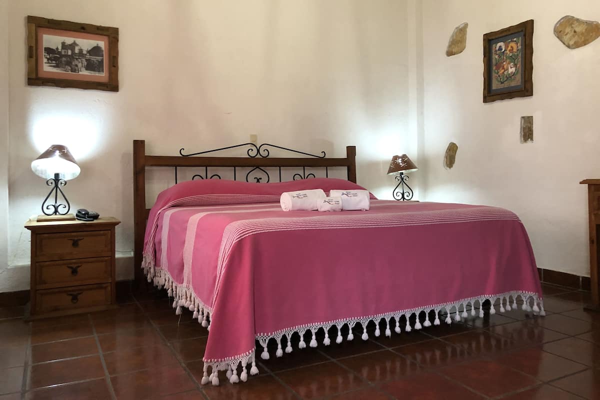 Una cama bien hecha con una colcha rosa y flecos blancos, una mesita de noche de madera con una lámpara, y un pequeño aparador de madera.