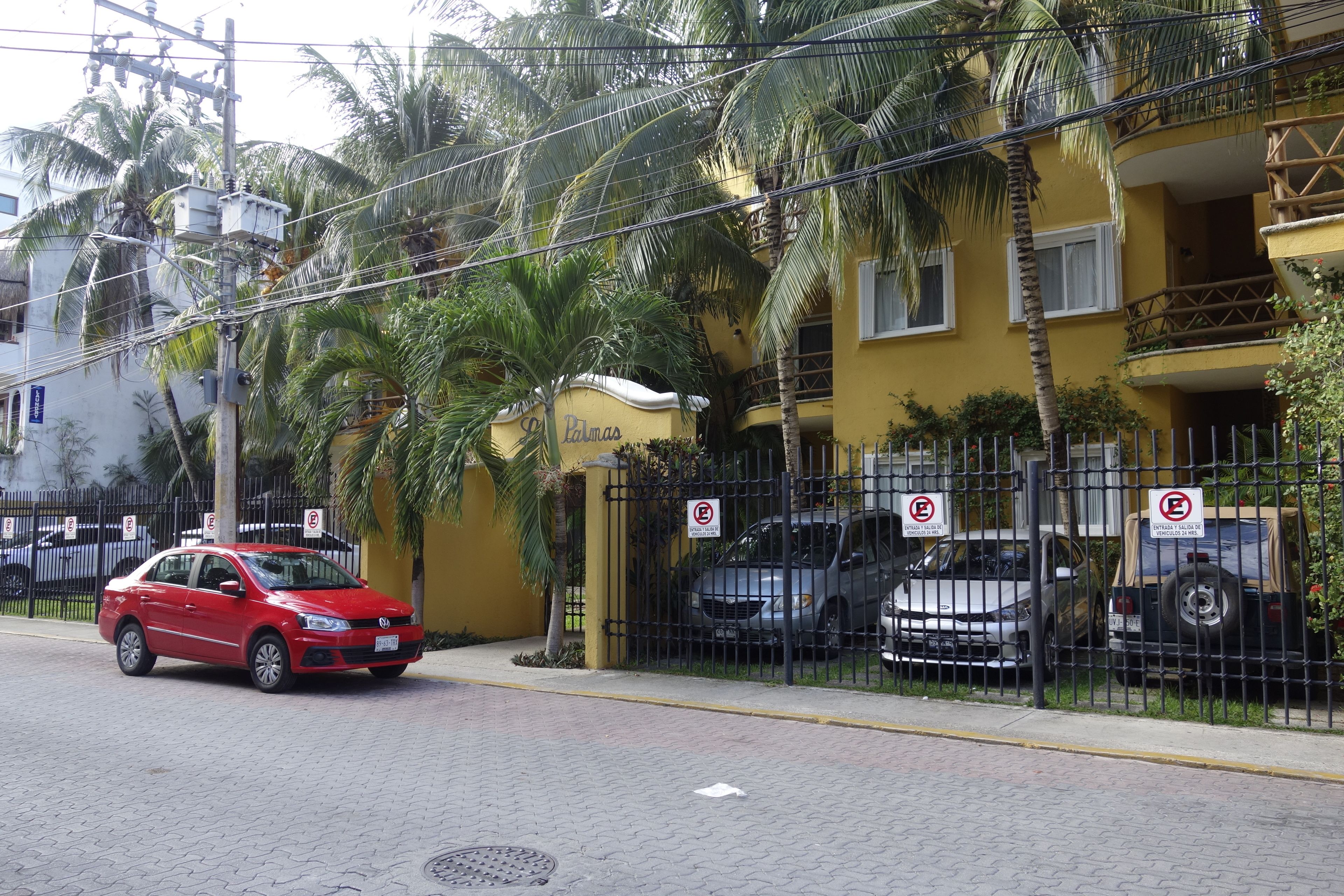Une voiture rouge est garée sur une rue pavée, avec un bâtiment jaune et une clôture noire sur laquelle on peut voir des panneaux indiquant « Stationnement interdit » en arrière-plan.