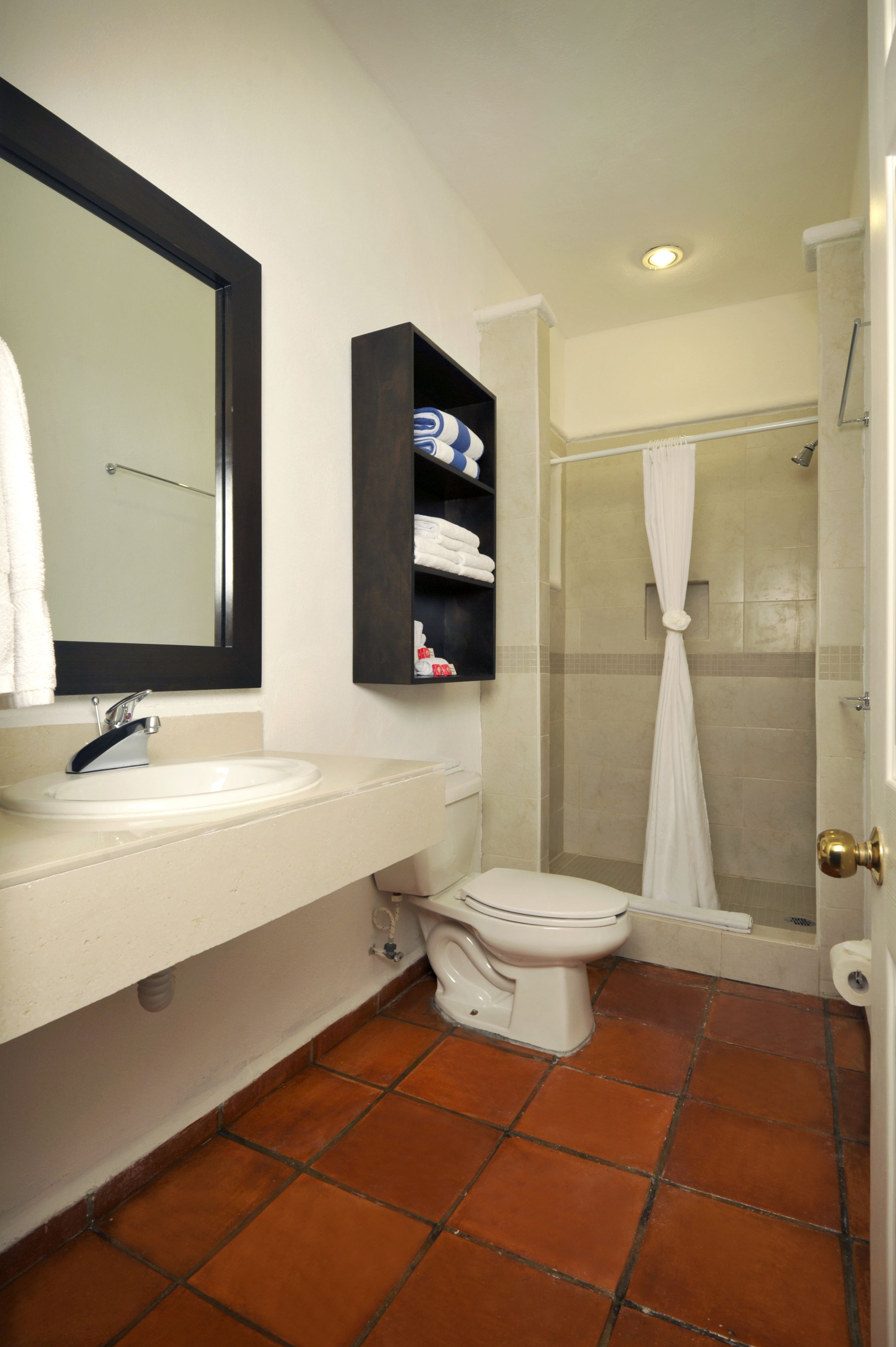 Un baño con un lavamanos blanco, un espejo, un inodoro, una ducha con cortina blanca y un estante con artículos de aseo.