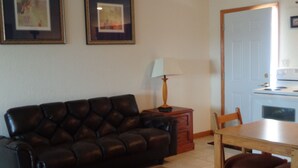 Una sala de estar con un sofá de cuero marrón oscuro, una mesa de madera con dos sillas, una lámpara y un tapete colorido. Hay cuadros enmarcados en la pared y se ve un área de cocina a través de una puerta abierta.