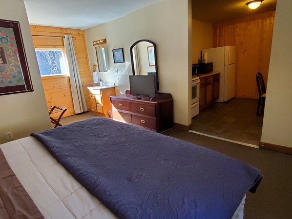 A bedroom with a bed, dresser, television, and a kitchenette in the background. - Kenosha Lodge (Grant)