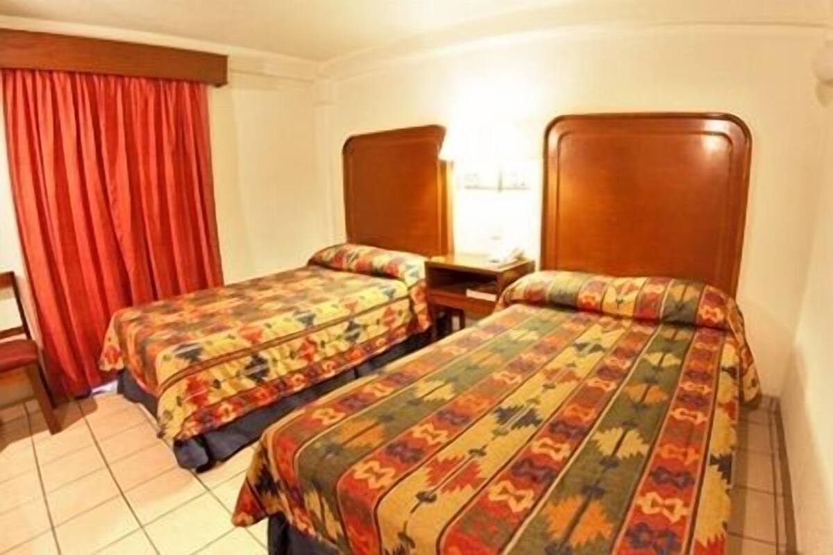 Una habitación de hotel con dos camas individuales, cabeceros de madera, una cortina roja y piso de baldosas.