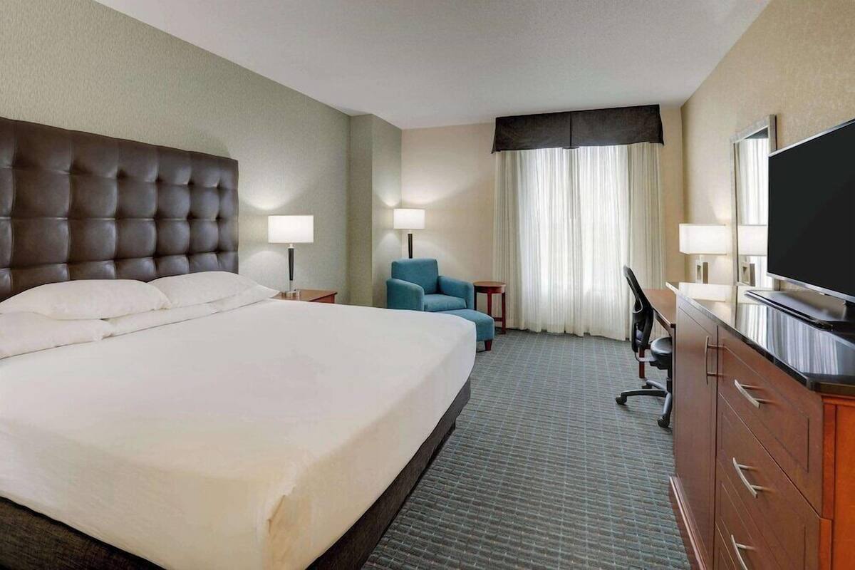 Ein Hotelzimmer mit einem großen Bett, einem Schreibtisch, einem Sessel, einem Fernseher und einem fenster mit Vorhängen.