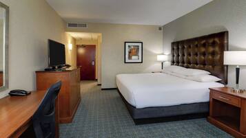 Quarto de hotel com cama, escrivaninha, cadeira, televisão e um quadro pendurado na parede.