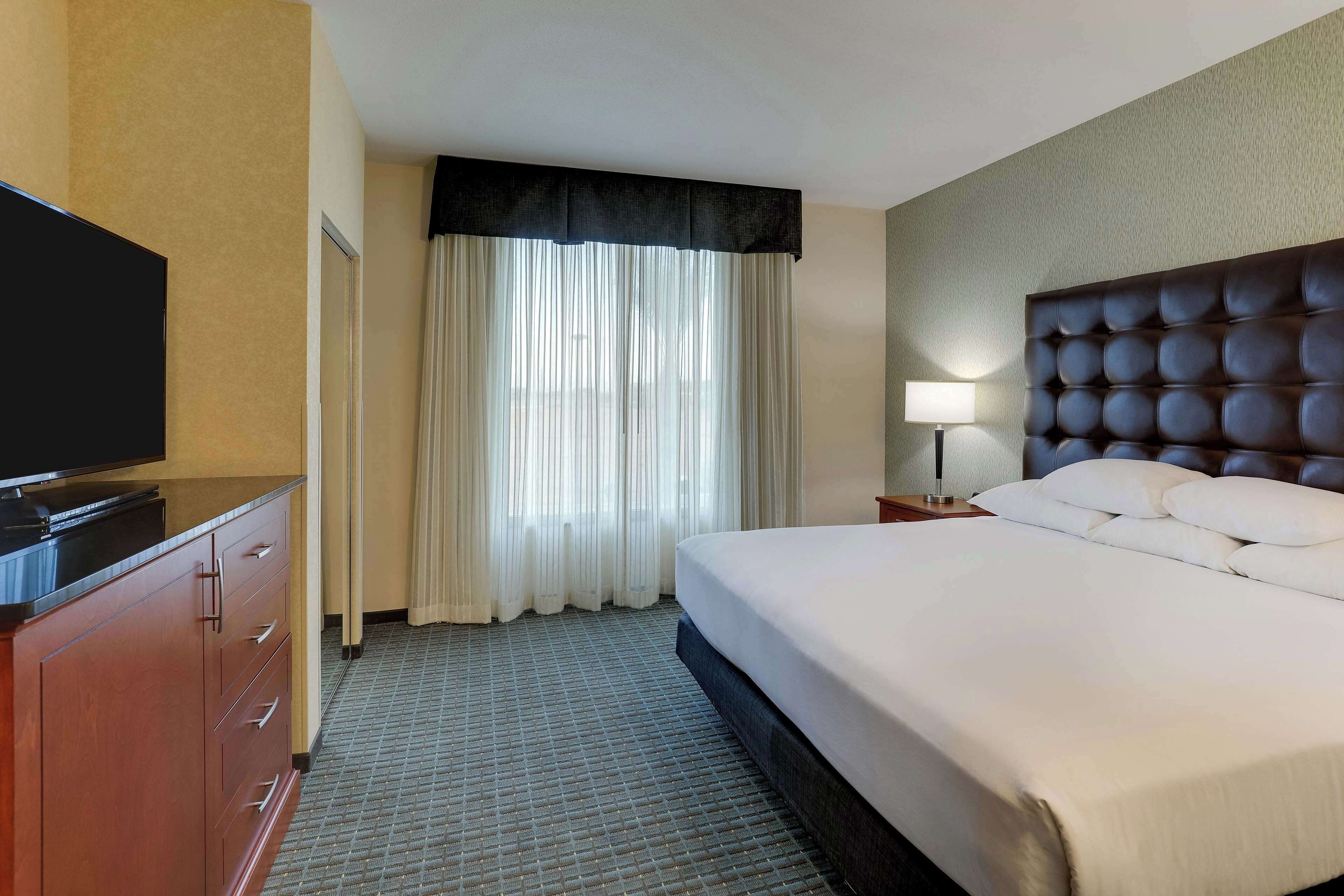 A hotel room with a large bed, a flat-screen TV on a wooden cabinet, a nightstand with a lamp, and a window with sheer curtains.