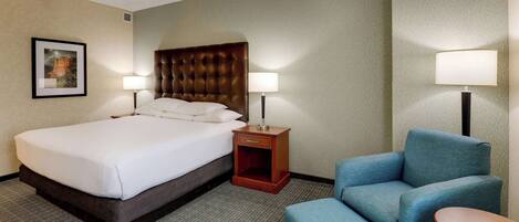 A hotel room with a large bed, two bedside tables with lamps, a blue armchair, and a small wooden table.