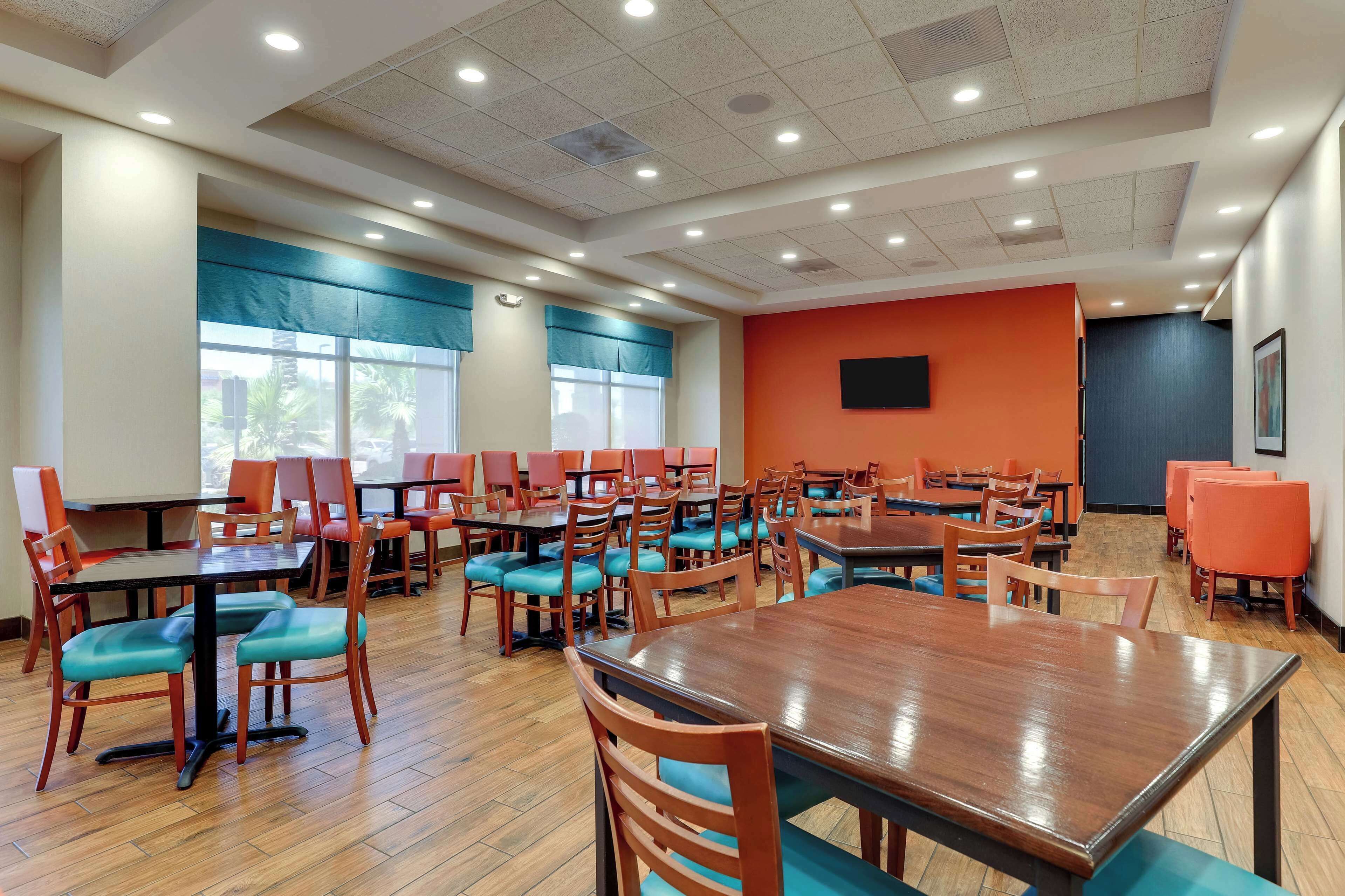 A spacious dining area with multiple tables and chairs, large windows, and a flat-screen TV mounted on the wall.