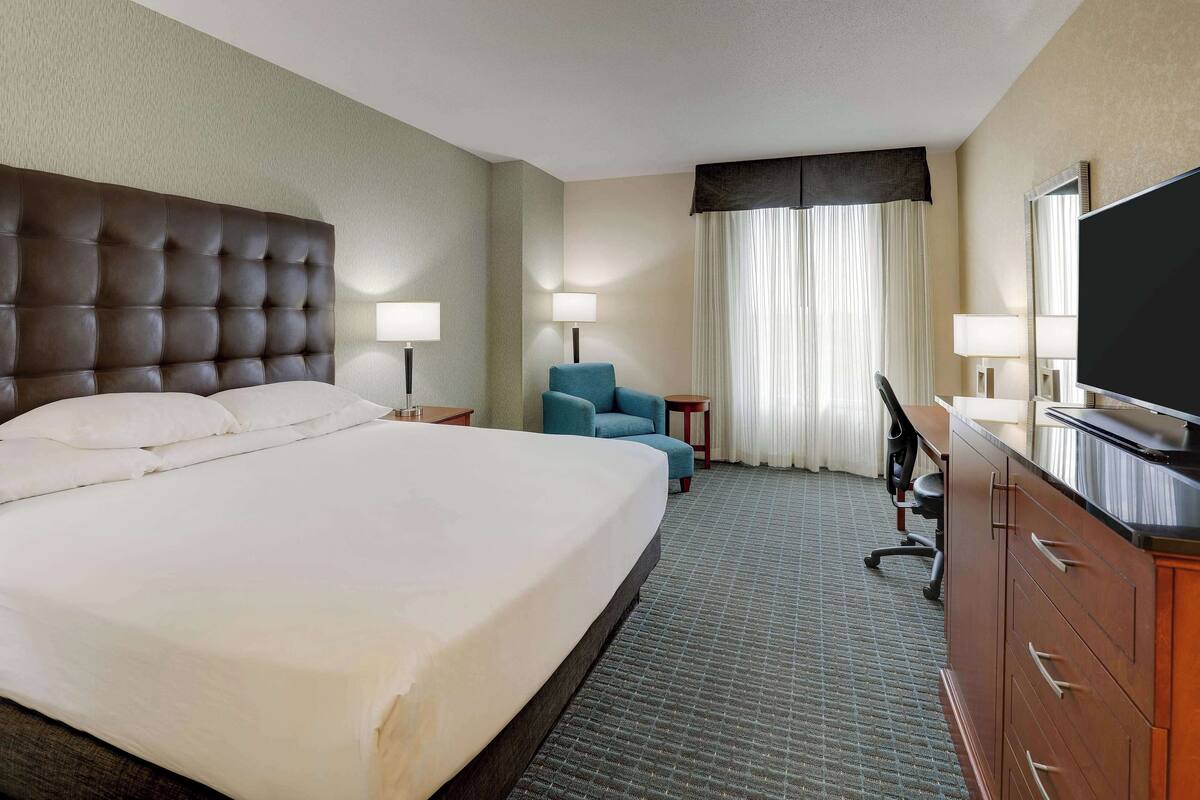 Ein Hotelzimmer mit einem großen Bett, einem Schreibtisch mit Stuhl, einem Fernseher und einem Fenster mit Vorhängen.