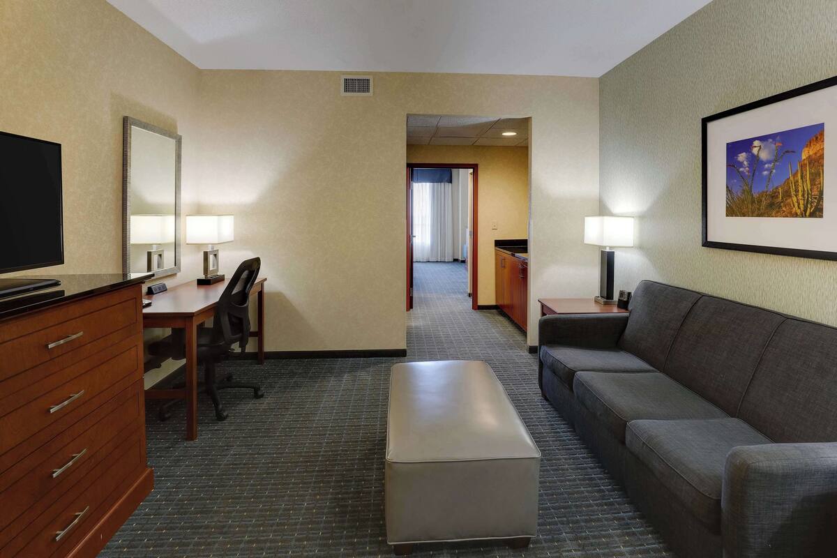 Ein Hotelzimmer mit Schreibtisch, Stuhl, Sofa, Hocker und Fernseher.