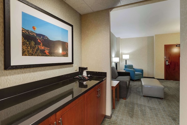 Un vestíbulo de hotel con un mostrador negro, un cuadro enmarcado de un globo aerostático, un sillón azul, un sofá gris y una puerta de madera.