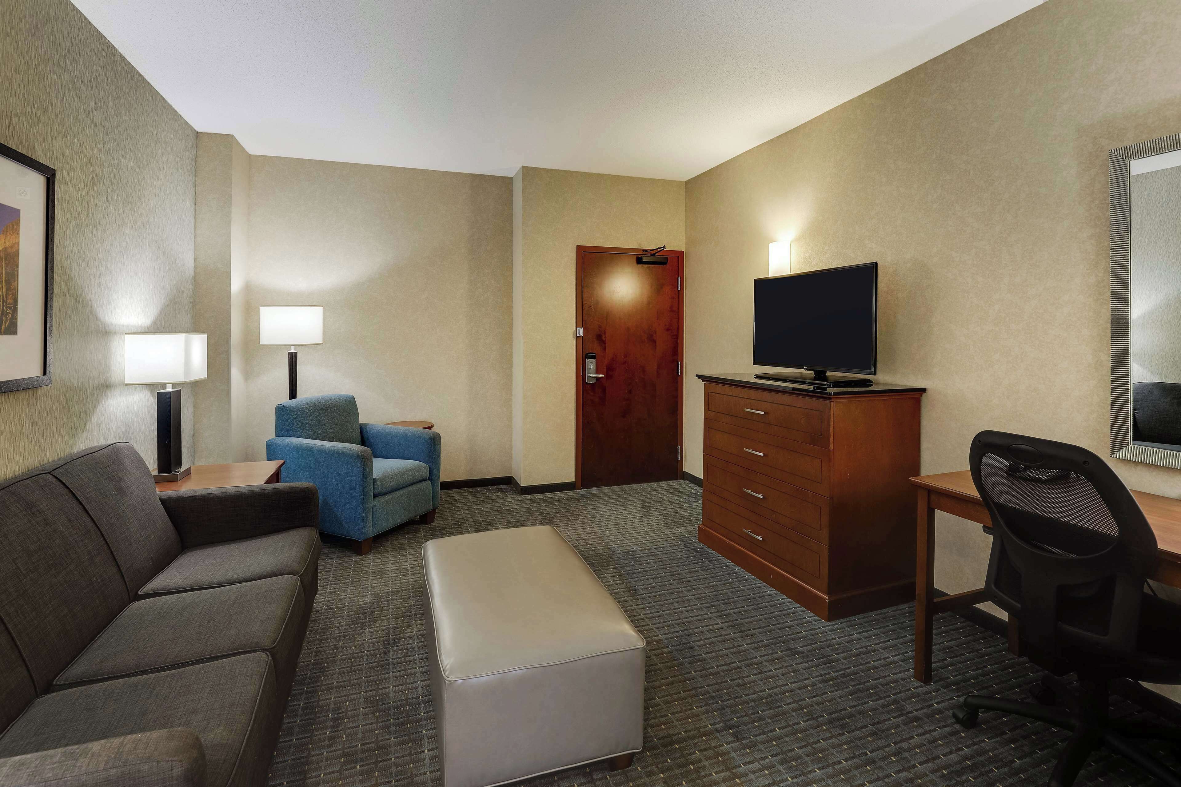 A hotel room with a sofa, a small blue chair, a desk with a computer, a television, a dresser, and a door.
